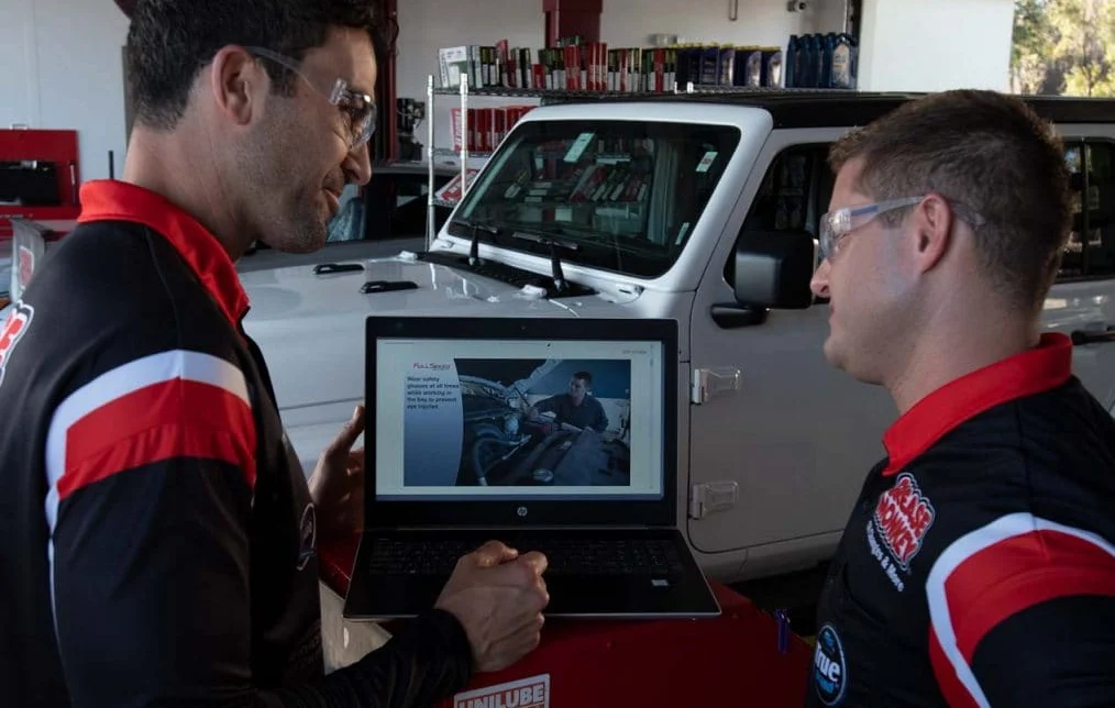 Two men in matching uniforms and safety glasses stand in a garage, looking at a laptop showing a car tutorial, with a white vehicle in the background.