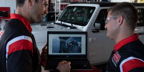 Two men in matching uniforms and safety glasses stand in a garage, looking at a laptop showing a car tutorial, with a white vehicle in the background.