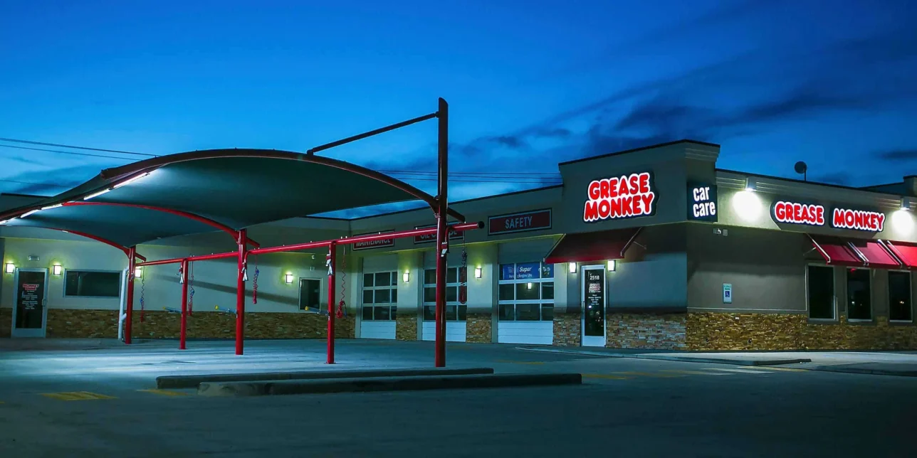 A Grease Monkey auto service shop with illuminated signs at dusk, featuring a covered drive-thru area and an empty parking lot.