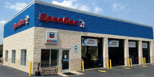 A SpeeDee Oil Change and Auto Service building with blue and tan exterior, signs for open Sunday and now hiring, and three service bays with yellow bollards.