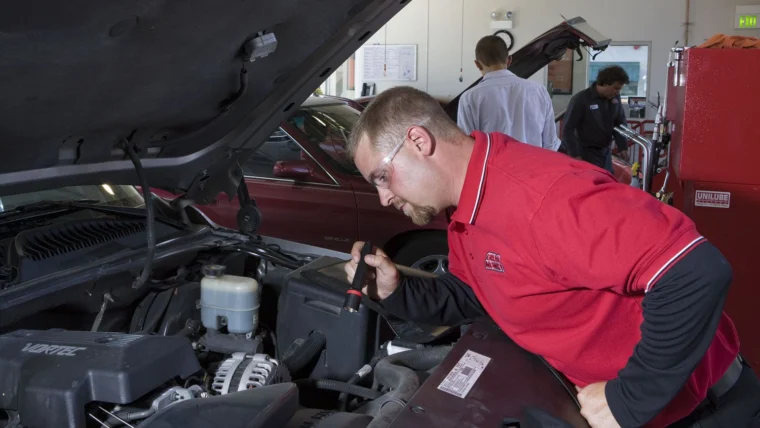 A man in safety glasses and a red shirt inspects a car engine with a flashlight in an auto repair shop, while three other people work in the background.