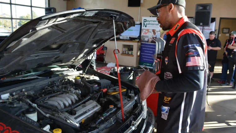 A mechanic in uniform inspects a car engine with the hood open, holding a cloth and dipstick inside an automotive service shop.