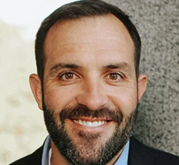 Jim Boswell with short dark hair and a beard, smiling, wearing a blue shirt and dark jacket, standing in front of a stone wall.