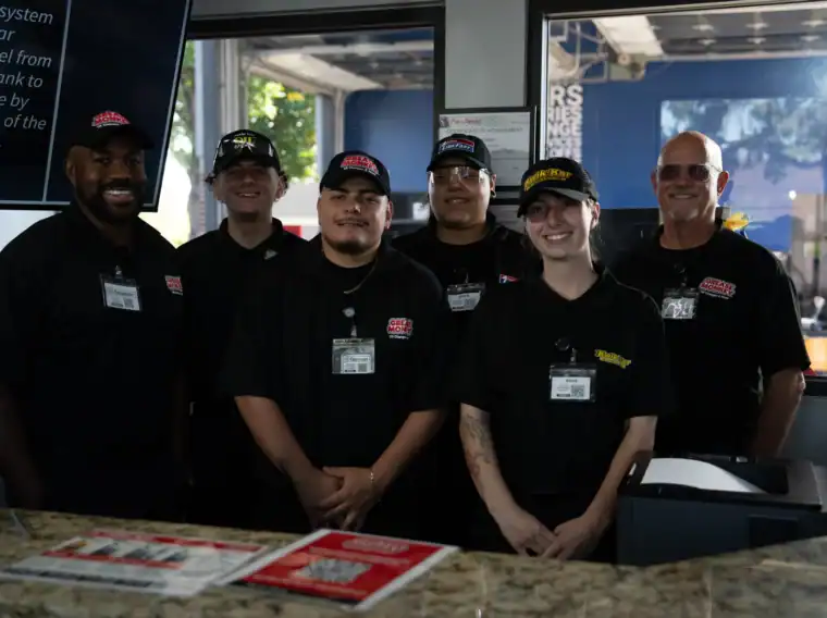 Six employees in black uniforms and hats stand behind a counter in an automotive service shop, smiling at the camera.