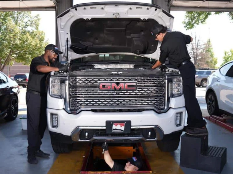 Three mechanics service a white GMC truck in a garage; two work under the hood, while one works underneath the vehicle in a maintenance pit.