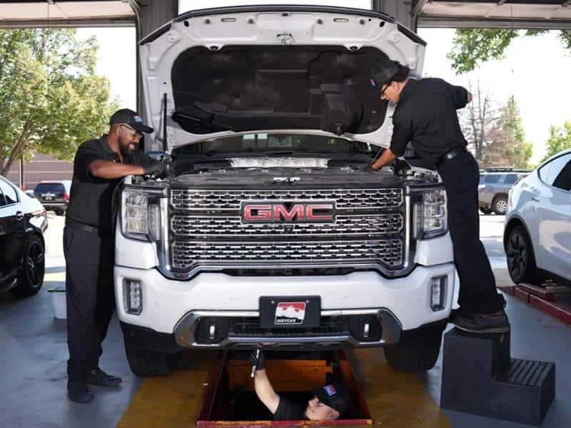 Three mechanics service a white GMC truck in a garage; two work under the hood, while one works underneath the vehicle in a maintenance pit.