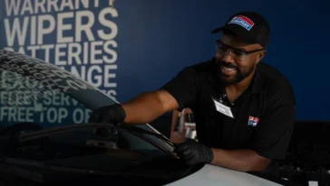 A worker wearing gloves and a cap is cleaning a car windshield inside an automotive service center.