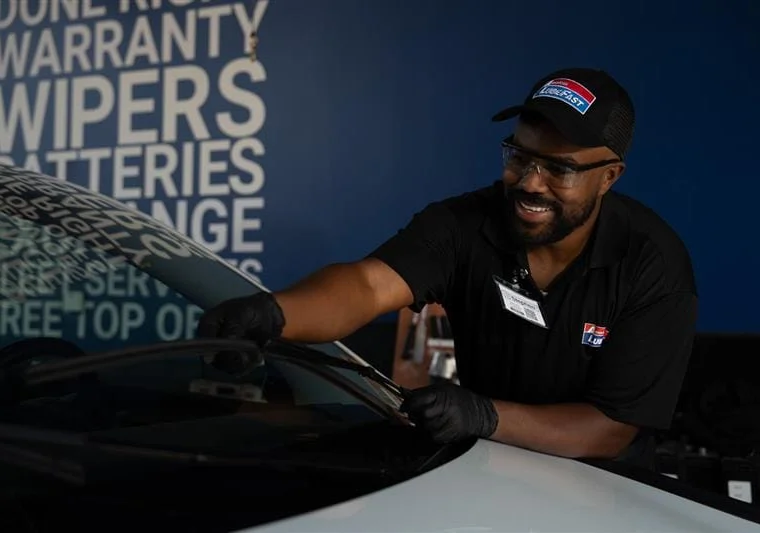 A worker wearing gloves and a cap is cleaning a car windshield inside an automotive service center.