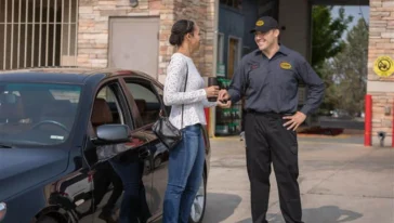 A woman stands by a black car handing keys to a uniformed employee outside an automotive service center.