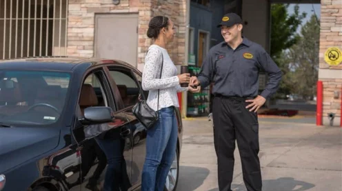 A woman stands by a black car handing keys to a uniformed employee outside an automotive service center.