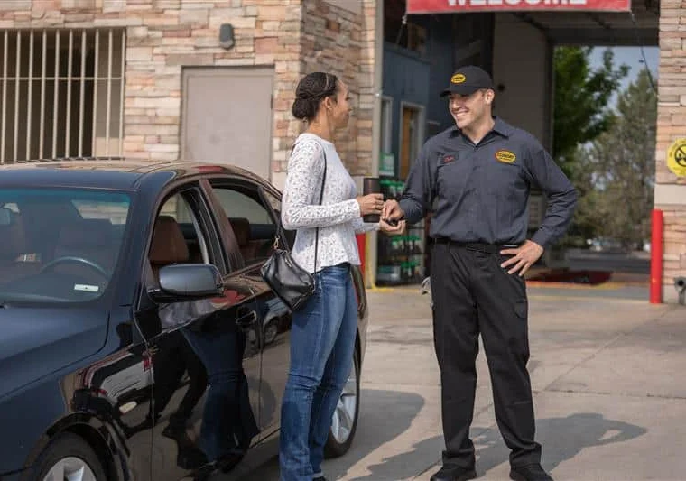 A woman stands by a black car handing keys to a uniformed employee outside an automotive service center.