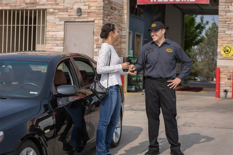 A woman stands by a black car handing keys to a uniformed employee outside an automotive service center.