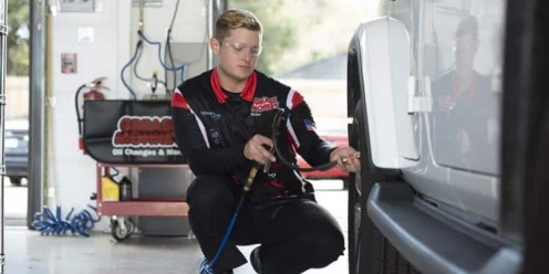A mechanic in uniform uses an air hose to check or inflate the tire of a white SUV in a well-lit garage.