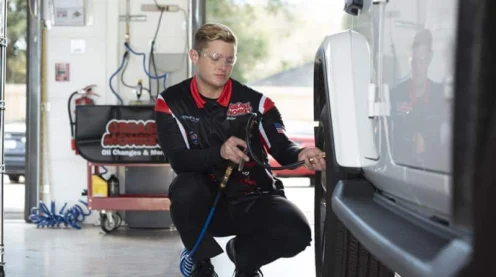 A mechanic in uniform uses an air hose to check or inflate the tire of a white SUV in a well-lit garage.