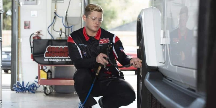 A mechanic in uniform uses an air hose to check or inflate the tire of a white SUV in a well-lit garage.