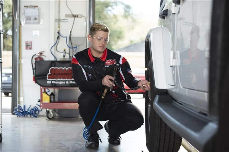 A mechanic in uniform uses an air hose to check or inflate the tire of a white SUV in a well-lit garage.