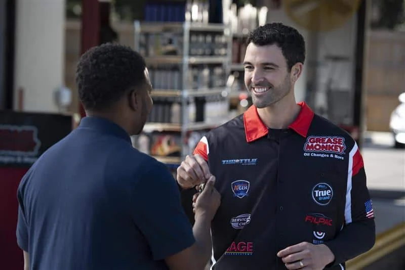Two men converse as one hands car keys to the other, who is wearing a "Grease Monkey" auto service uniform in a garage setting.