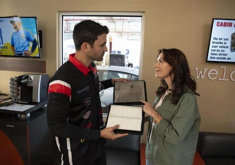 A mechanic shows a woman two cabin air filters, one clean and one dirty, inside an auto service waiting area.