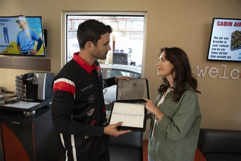 A mechanic shows a woman two cabin air filters, one clean and one dirty, inside an auto service waiting area.