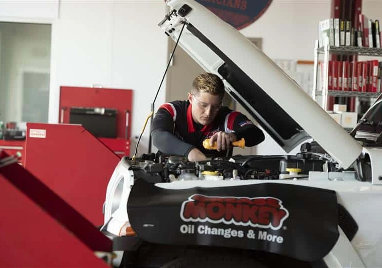 A mechanic works under the hood of a vehicle in a garage, with a "Monkey Oil Changes & More" cover draped over the front.