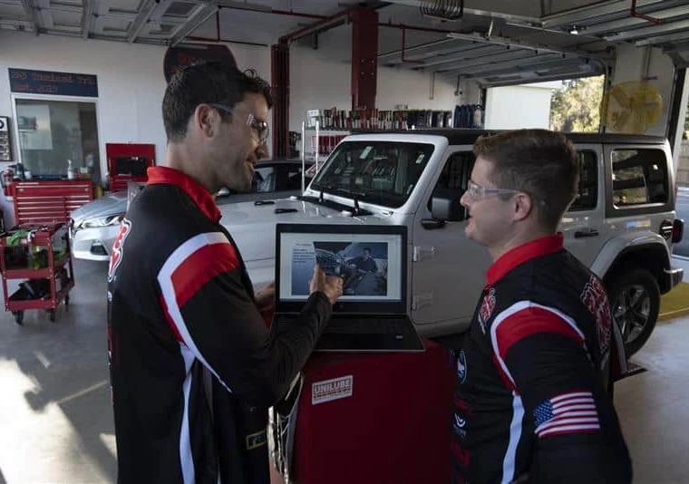 Two men in matching uniforms discuss information displayed on a laptop in an automotive garage, with vehicles and tools visible in the background.