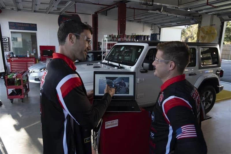 Two men in matching uniforms discuss information displayed on a laptop in an automotive garage, with vehicles and tools visible in the background.