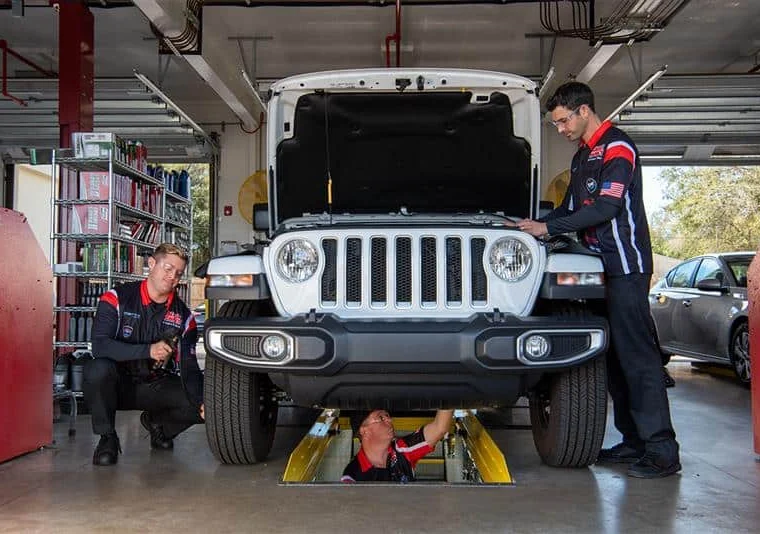 Three mechanics inspect and work on a white SUV in a garage; one is beneath the vehicle while two others stand and kneel beside it.