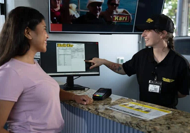 A customer smiles at a staff member behind a counter as the staff member points to information on a computer screen. A payment terminal and brochure are on the counter.