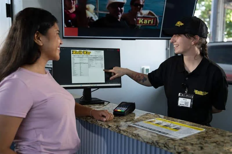 A customer smiles at a staff member behind a counter as the staff member points to information on a computer screen. A payment terminal and brochure are on the counter.
