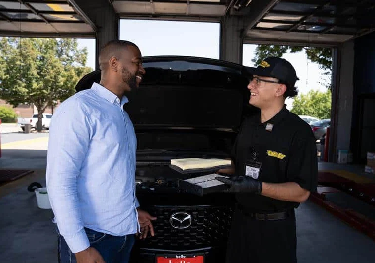A customer and an auto technician stand in front of a Mazda vehicle with its hood open, talking and smiling inside an automotive service garage.