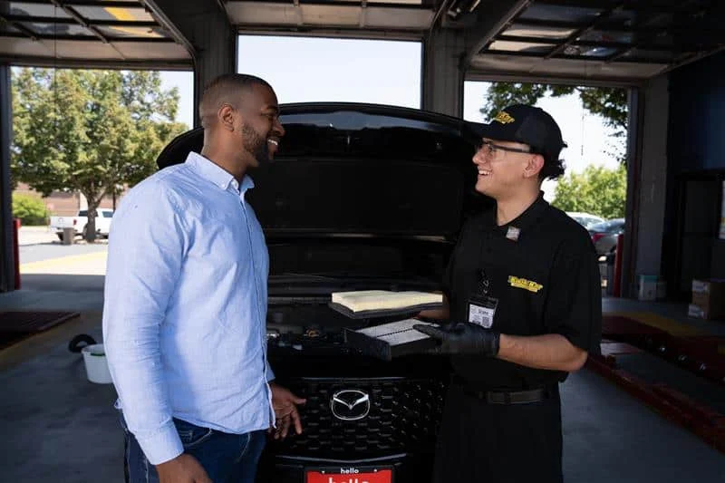 A customer and an auto technician stand in front of a Mazda vehicle with its hood open, talking and smiling inside an automotive service garage.