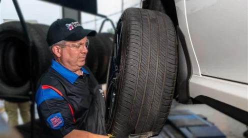 An auto mechanic wearing gloves and safety glasses installs a tire onto a raised vehicle in a service garage.