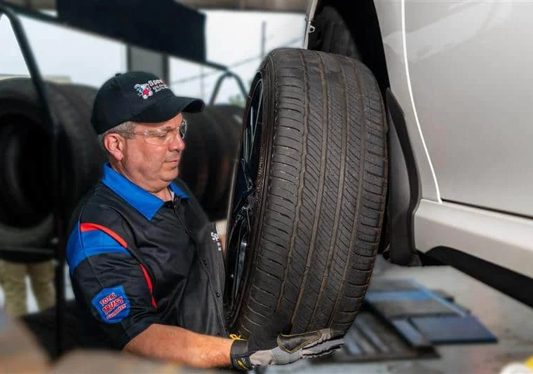 An auto mechanic wearing gloves and safety glasses installs a tire onto a raised vehicle in a service garage.