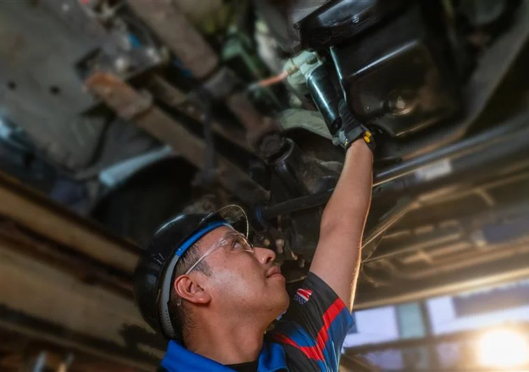 A mechanic wearing safety glasses and a hard hat inspects or repairs the underside of a vehicle in a workshop setting.