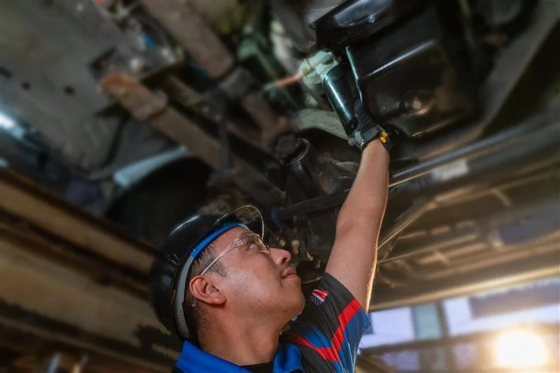 A mechanic wearing safety glasses and a hard hat inspects or repairs the underside of a vehicle in a workshop setting.