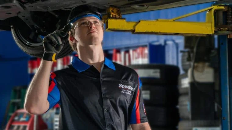 A mechanic wearing safety glasses and gloves inspects the underside of a vehicle on a lift in an auto repair shop.