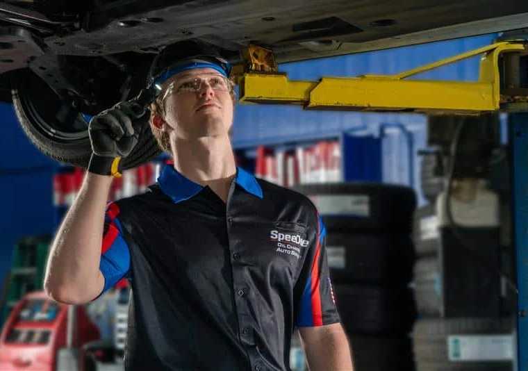 A mechanic wearing safety glasses and gloves inspects the underside of a vehicle on a lift in an auto repair shop.