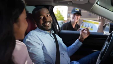 A man in the driver's seat smiles at a woman beside him while a fast food worker hands him a menu through the car window.
