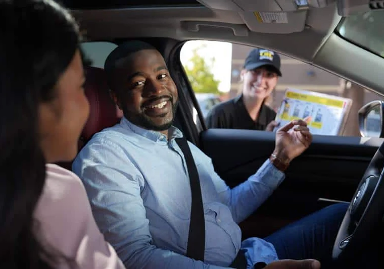 A man in the driver's seat smiles at a woman beside him while a fast food worker hands him a menu through the car window.