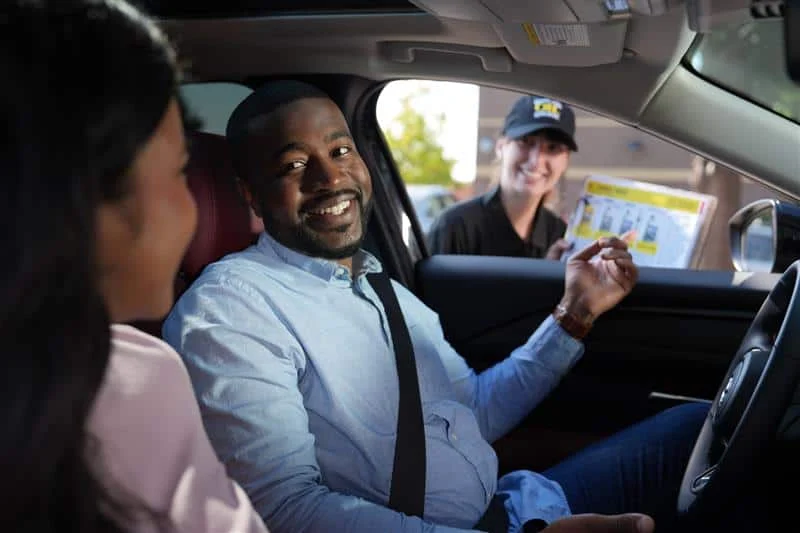 A man in the driver's seat smiles at a woman beside him while a fast food worker hands him a menu through the car window.