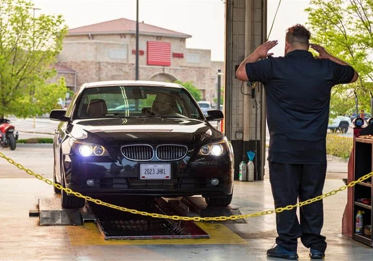 A black BMW sedan enters a vehicle inspection station as an attendant signals with both hands raised.