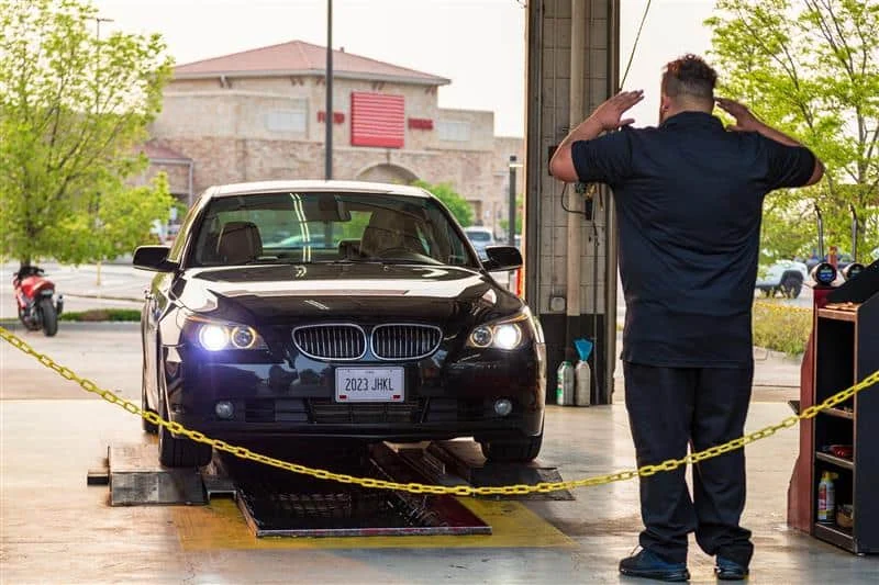 A black BMW sedan enters a vehicle inspection station as an attendant signals with both hands raised.