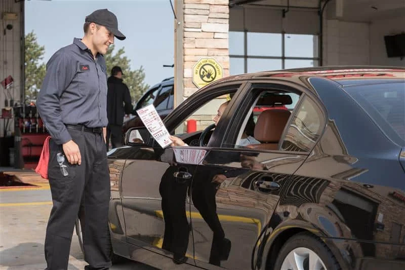 A mechanic in uniform hands a paper to a driver sitting in a black car at an auto service center.
