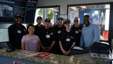 A group of employees in uniforms and two people in casual clothes stand behind a service counter, smiling for a group photo inside an automotive shop.