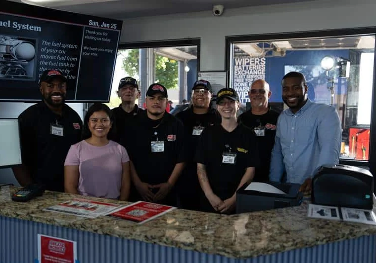 A group of employees in uniforms and two people in casual clothes stand behind a service counter, smiling for a group photo inside an automotive shop.