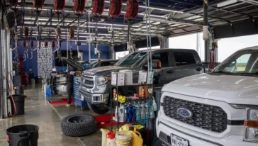 Vehicles inside an auto repair shop, with tools, oil containers, and equipment visible; one vehicle has its hood open for maintenance.