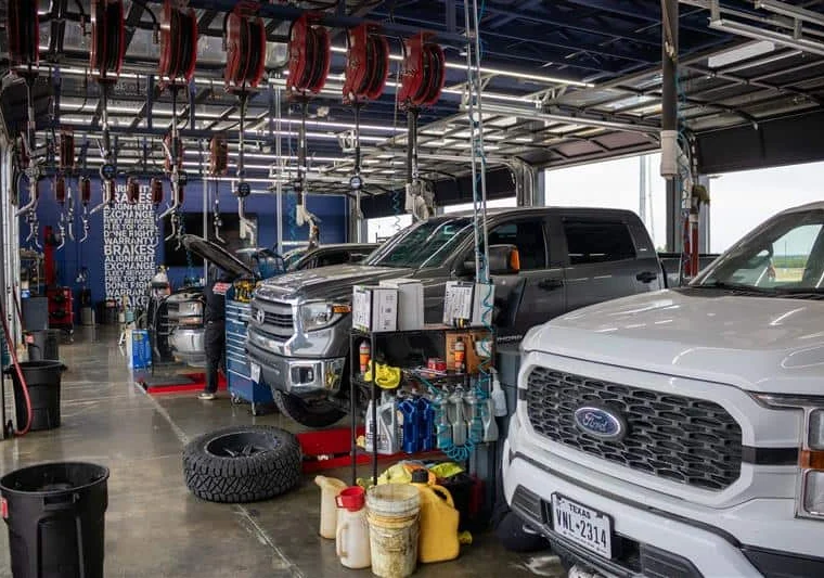 Vehicles inside an auto repair shop, with tools, oil containers, and equipment visible; one vehicle has its hood open for maintenance.