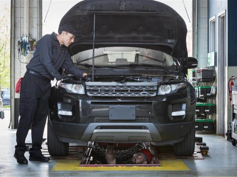 A mechanic inspects the engine of a black SUV with its hood open, while another person works underneath the vehicle on a garage lift.