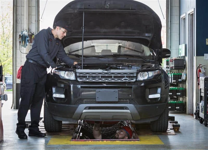 A mechanic inspects the engine of a black SUV with its hood open, while another person works underneath the vehicle on a garage lift.