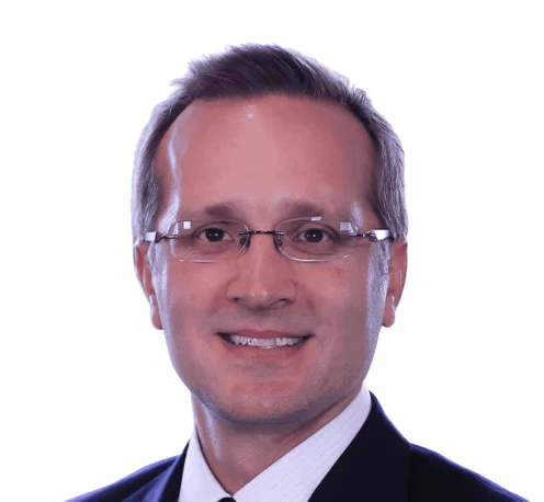 Brian Maciak wearing glasses, a dark suit, white shirt, and dark tie, smiling at the camera against a plain white background.
